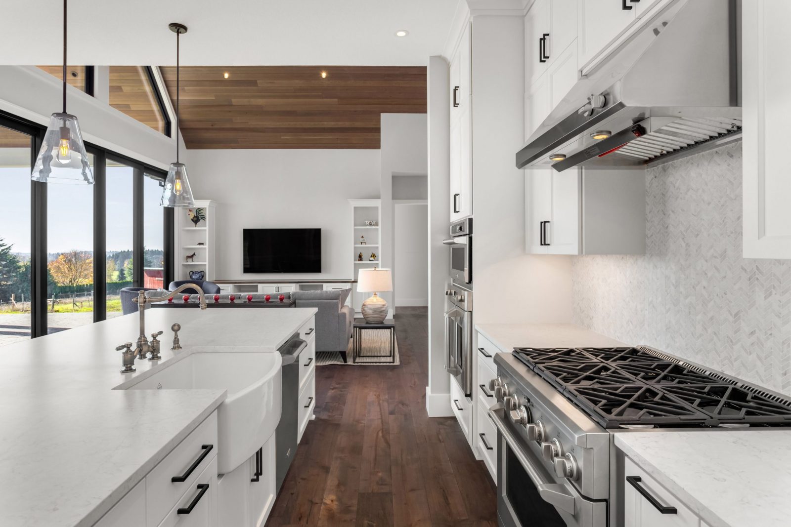 Beautiful kitchen and living room in new luxury home. Features large island with farmhouse sink, stainless steel appliances, gas range, and hardwood floors. Living room has vaulted ceilings and tv.